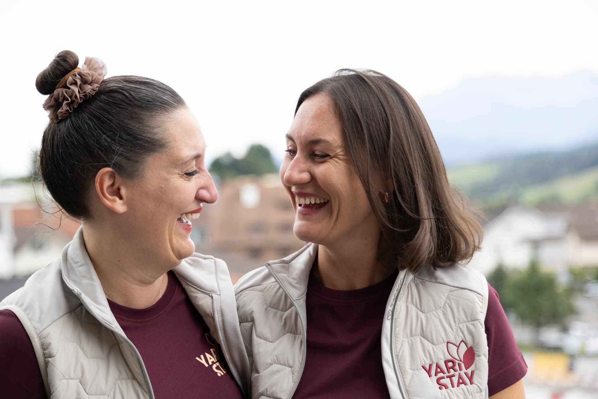 Sibille und Marianne, die Gründerinnen der Yari Stay GmbH, in ihren Firmenkleidern lachend im Freien mit Berglandschaft im Hintergrund